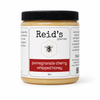 A 9oz jar of Reid's Gourmet Pomegranate Cherry Whipped Honey on a clean white background. The jar has a black ribbed lid and a white label with a red banner displaying the product name.